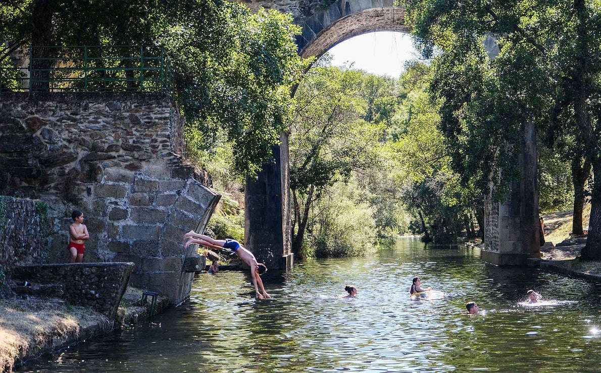 Un joven se lanza de cabeza al agua en la piscina natural de Perales del Puerto, con el puente al fondo.